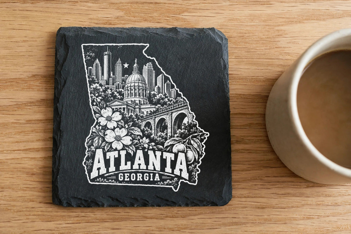 A square black slate coaster featuring an engraved white illustration of Atlanta landmarks inside a state outline.