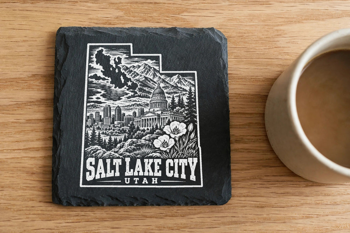 A square slate coaster engraved with a Salt Lake City skyline graphic sits on a wooden table.