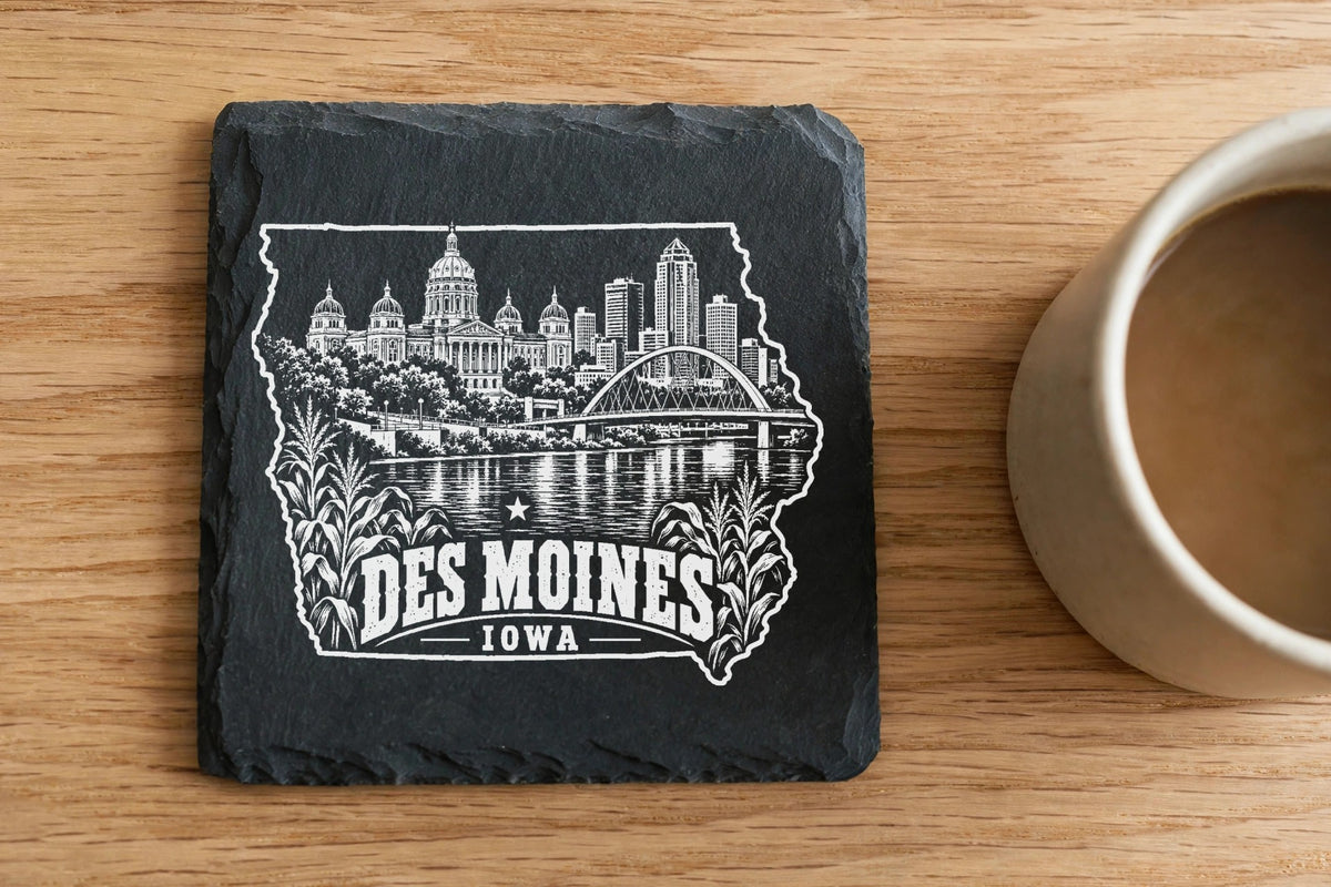 A square black slate coaster engraved with a Des Moines skyline illustration rests on a wooden table beside a coffee mug.