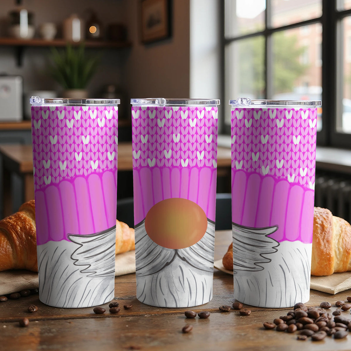 Three tumblers with pink knit and gnome designs on a wooden table with croissants.