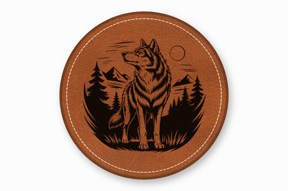A round brown leather coaster features a black laser-engraved design of a wolf in a forest landscape.