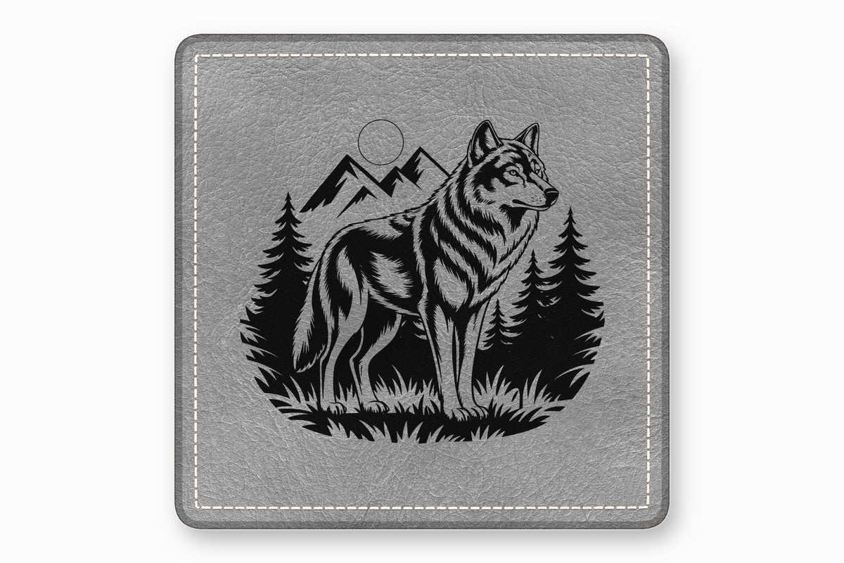 A square grey leather coaster featuring a black engraved design of a wolf, mountains, and pine trees.