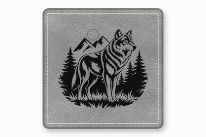 A square grey leather coaster featuring a black engraved design of a wolf, mountains, and pine trees.