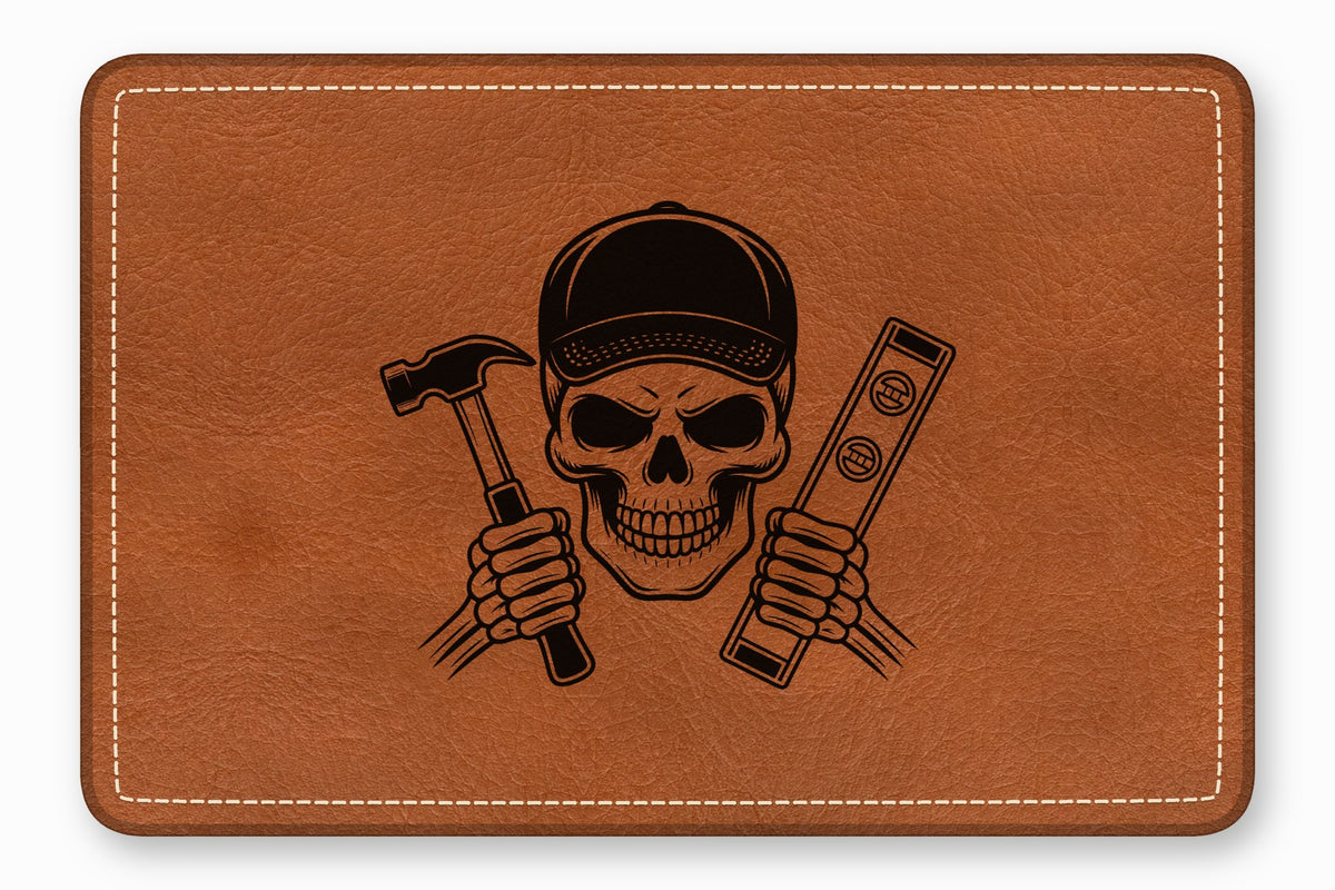 A brown leather patch with white stitched edges features a black engraved skull wearing a cap and holding a hammer and a level.