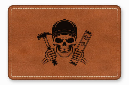 A brown leather patch with white stitched edges features a black engraved skull wearing a cap and holding a hammer and a level.
