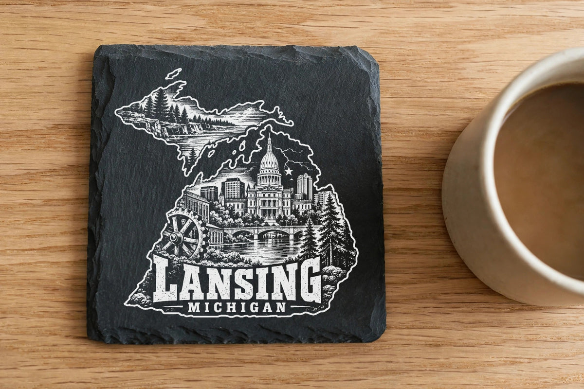 Square slate coaster engraved with a map of Michigan, Lansing city landmarks, and text next to a coffee mug on a wooden table.