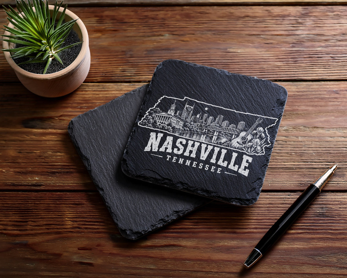 Two square black slate coasters engraved with the Nashville skyline resting on a wooden table.