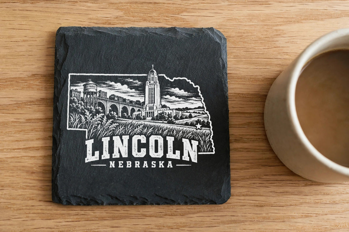 Square black slate coaster engraved with an outline of Nebraska featuring Lincoln landmarks next to a coffee mug.