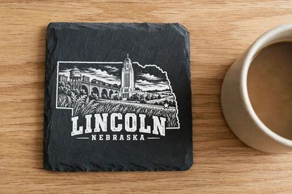Square black slate coaster engraved with an outline of Nebraska featuring Lincoln landmarks next to a coffee mug.