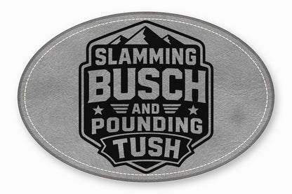 An oval grey leather patch featuring a black badge with mountains and the text Slamming Busch and Pounding Tush.