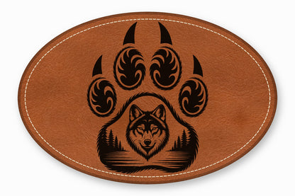 An oval brown leather patch with white stitched edges features a black bear paw graphic framing a wolf head and forest landscape.