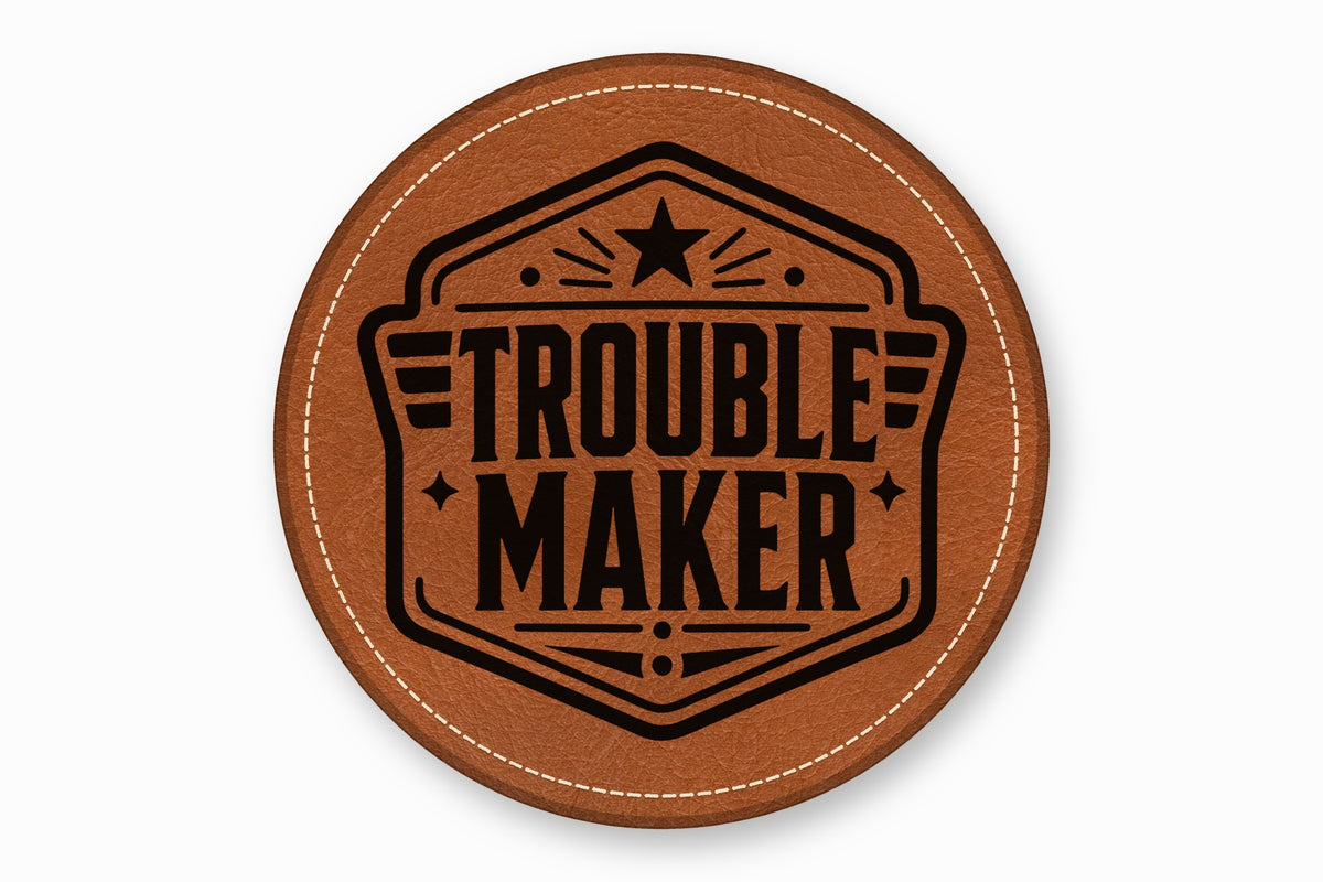 Round brown leather patch with white stitched border and black stamped text reading Trouble Maker.