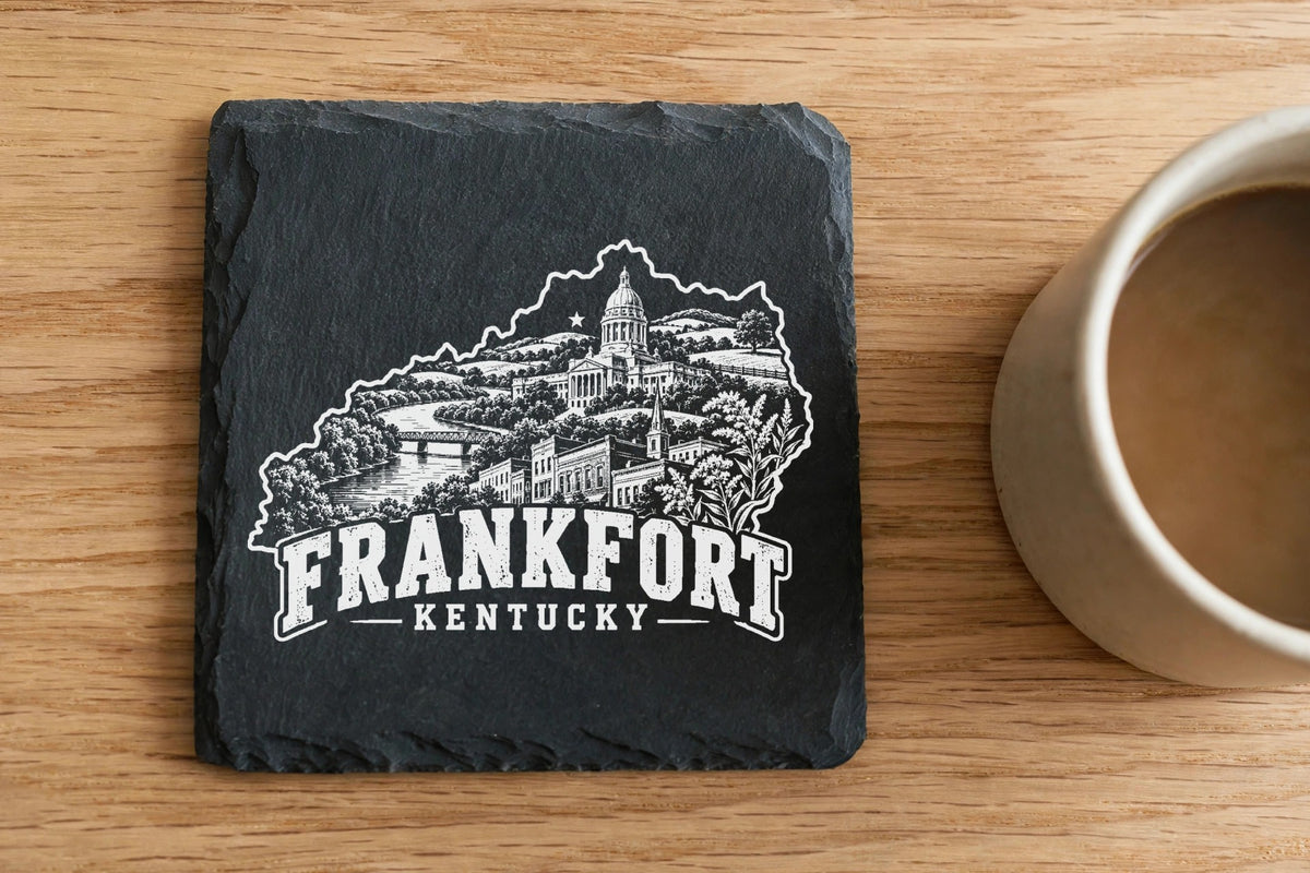 A square slate coaster with a white engraved map of Frankfort, Kentucky, on a wooden table.