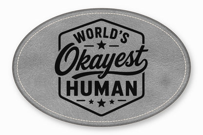 Oval gray leather patch with white stitching and the black text World's Okayest Human.