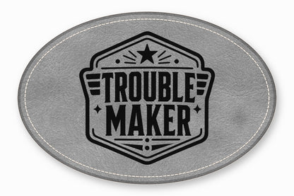 An oval grey leather patch with white stitched edges and a black Trouble Maker emblem.
