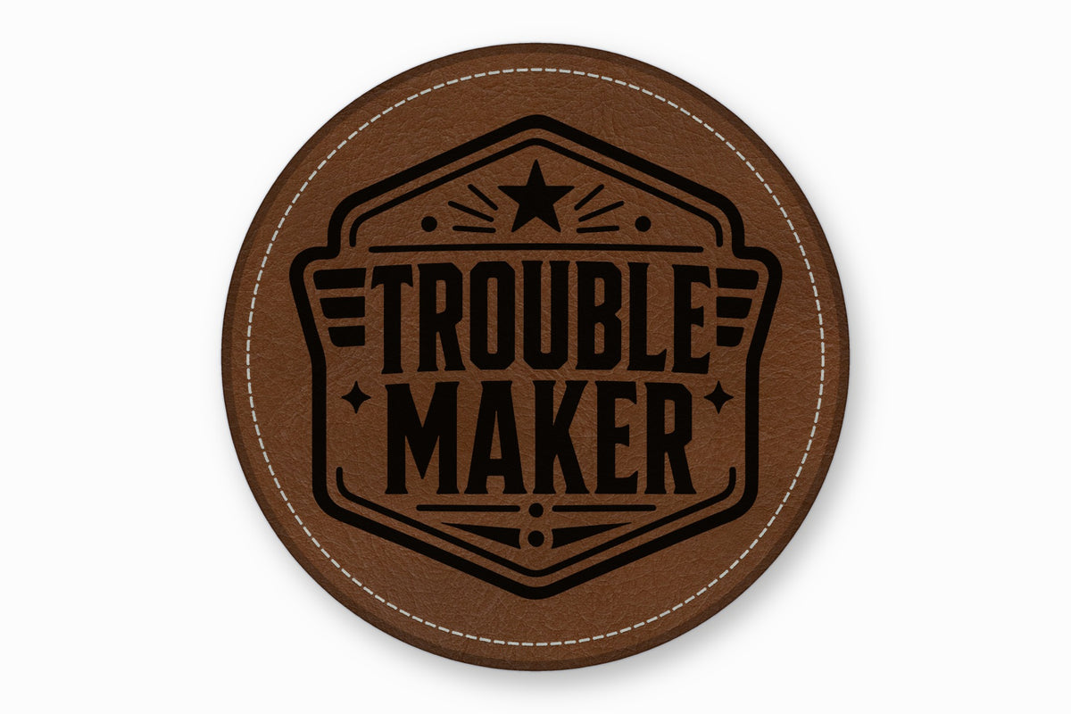 A brown round leather coaster features the black text Trouble Maker inside an illustrated emblem with stars.