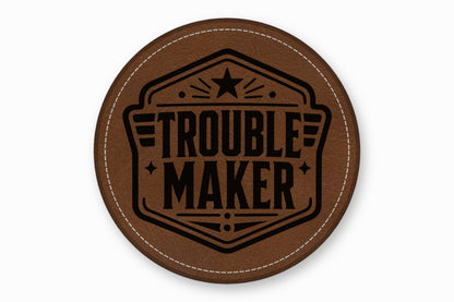 A brown round leather coaster features the black text Trouble Maker inside an illustrated emblem with stars.