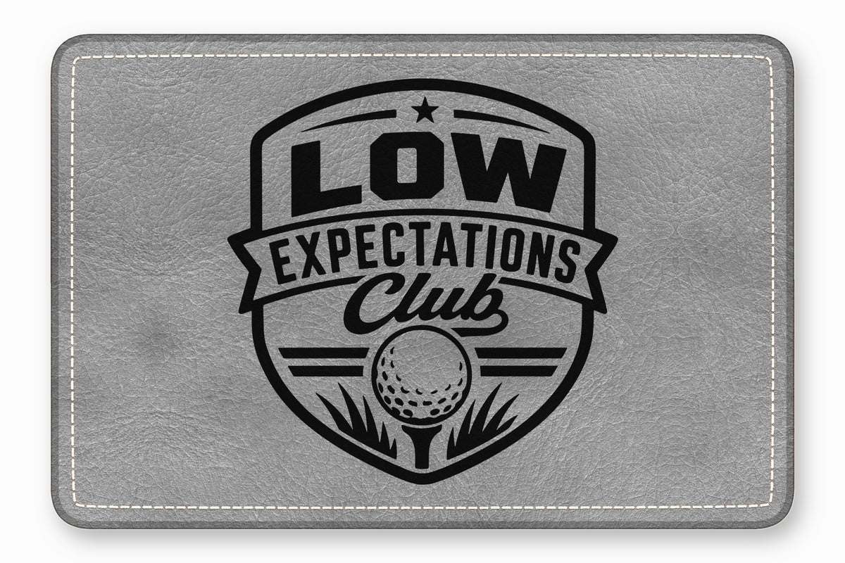 Gray leatherette patch engraved with a black Low Expectations Club golf crest.