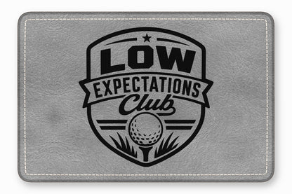 Gray leatherette patch engraved with a black Low Expectations Club golf crest.