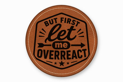 Round brown leather coaster with white stitched edges, featuring the black text BUT FIRST LET ME OVERREACT.