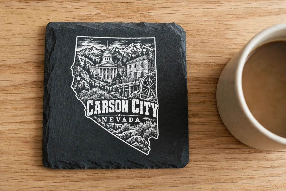 A square slate coaster engraved with a Carson City, Nevada map design sits on a wooden table beside a coffee mug.