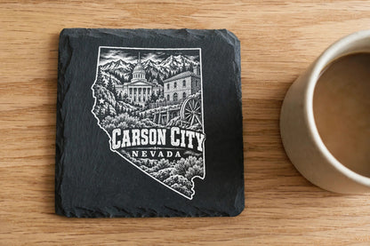 A square slate coaster engraved with a Carson City, Nevada map design sits on a wooden table beside a coffee mug.