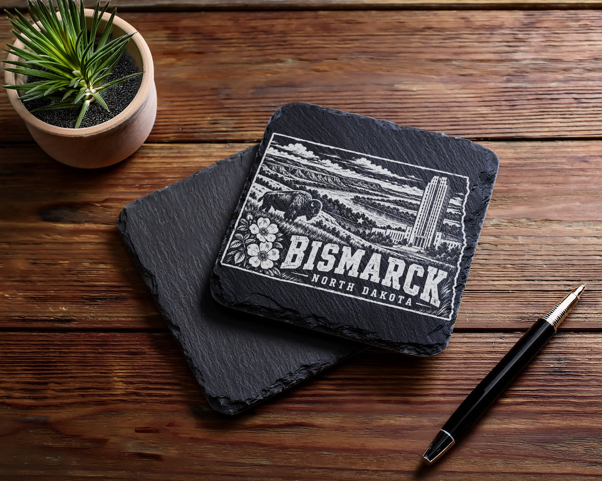 Two square black slate coasters on a wooden table, one engraved with a Bismarck, North Dakota skyline design.