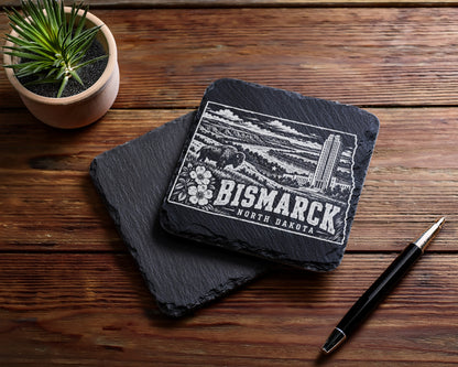 Two square black slate coasters on a wooden table, one engraved with a Bismarck, North Dakota skyline design.