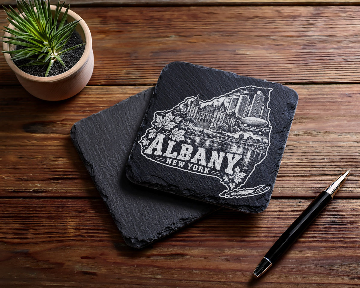 Two square slate coasters with an engraved Albany skyline design rest on a wooden table.