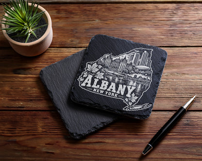 Two square slate coasters with an engraved Albany skyline design rest on a wooden table.