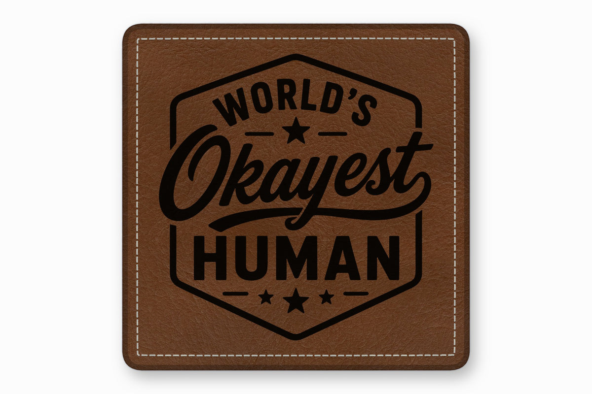 Square brown leather patch with white stitched borders and a black engraved text reading Worlds Okayest Human.