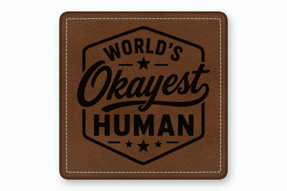 Square brown leather patch with white stitched borders and a black engraved text reading Worlds Okayest Human.