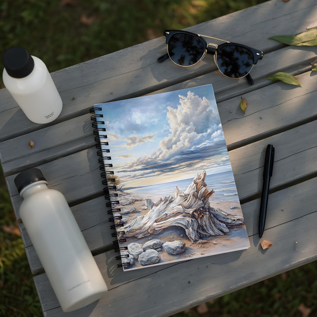 Spiral bound notebook with a beach landscape painting, a pen, and water bottles.