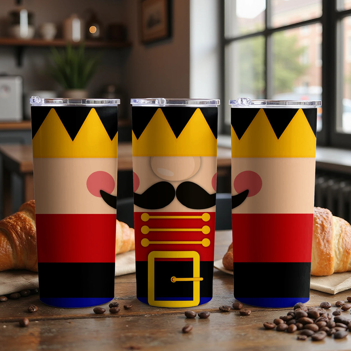 Three tumblers with nutcracker designs stand on a wooden table with croissants and coffee beans.