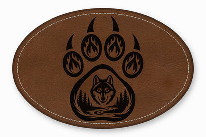Brown oval leather patch featuring a black bear paw print with a wolf and forest scene inside.