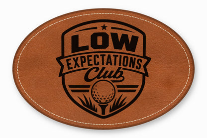 Oval brown leather patch with white stitched border, featuring a black Low Expectations Club golf logo.