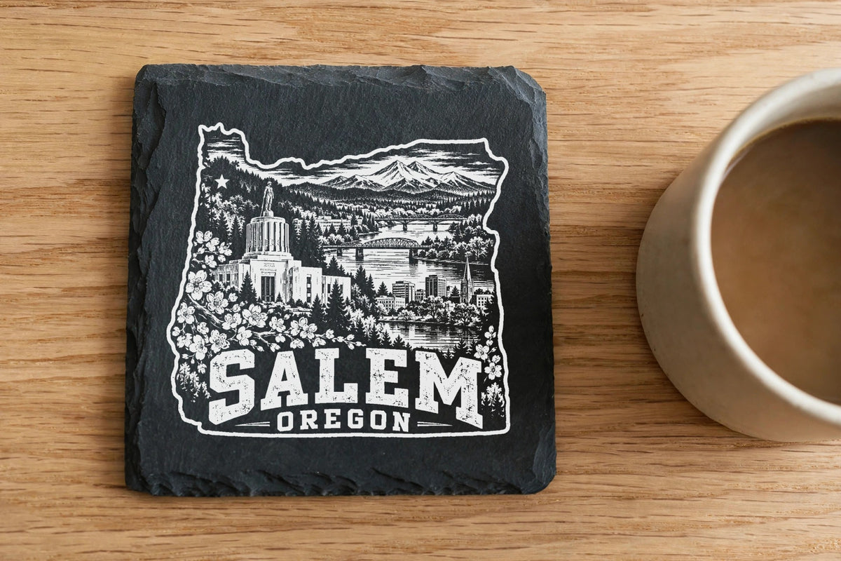 A square slate coaster with a white engraved map of Oregon and Salem text beside a coffee cup on a wooden table.