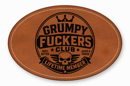 Oval brown leather patch with white stitched border and black debossed text reading Grumpy Fuckers Club.