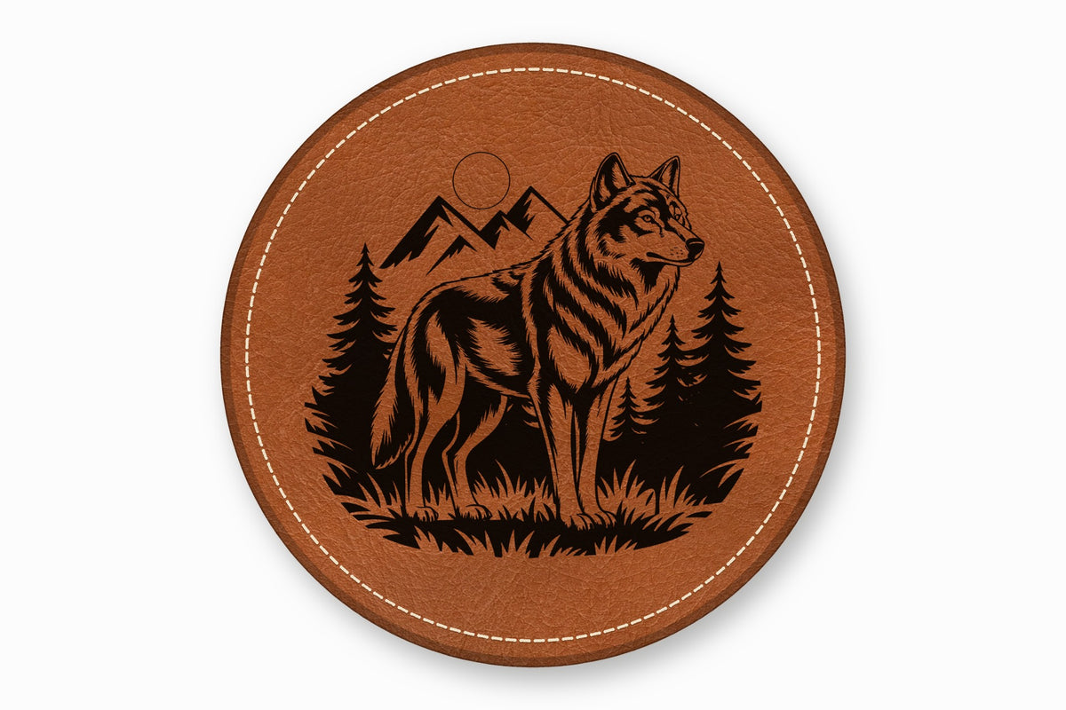 A round brown leather coaster with white stitched edges features a black laser-engraved wolf standing in a forest with mountains.