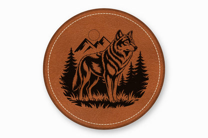 A round brown leather coaster with white stitched edges features a black laser-engraved wolf standing in a forest with mountains.