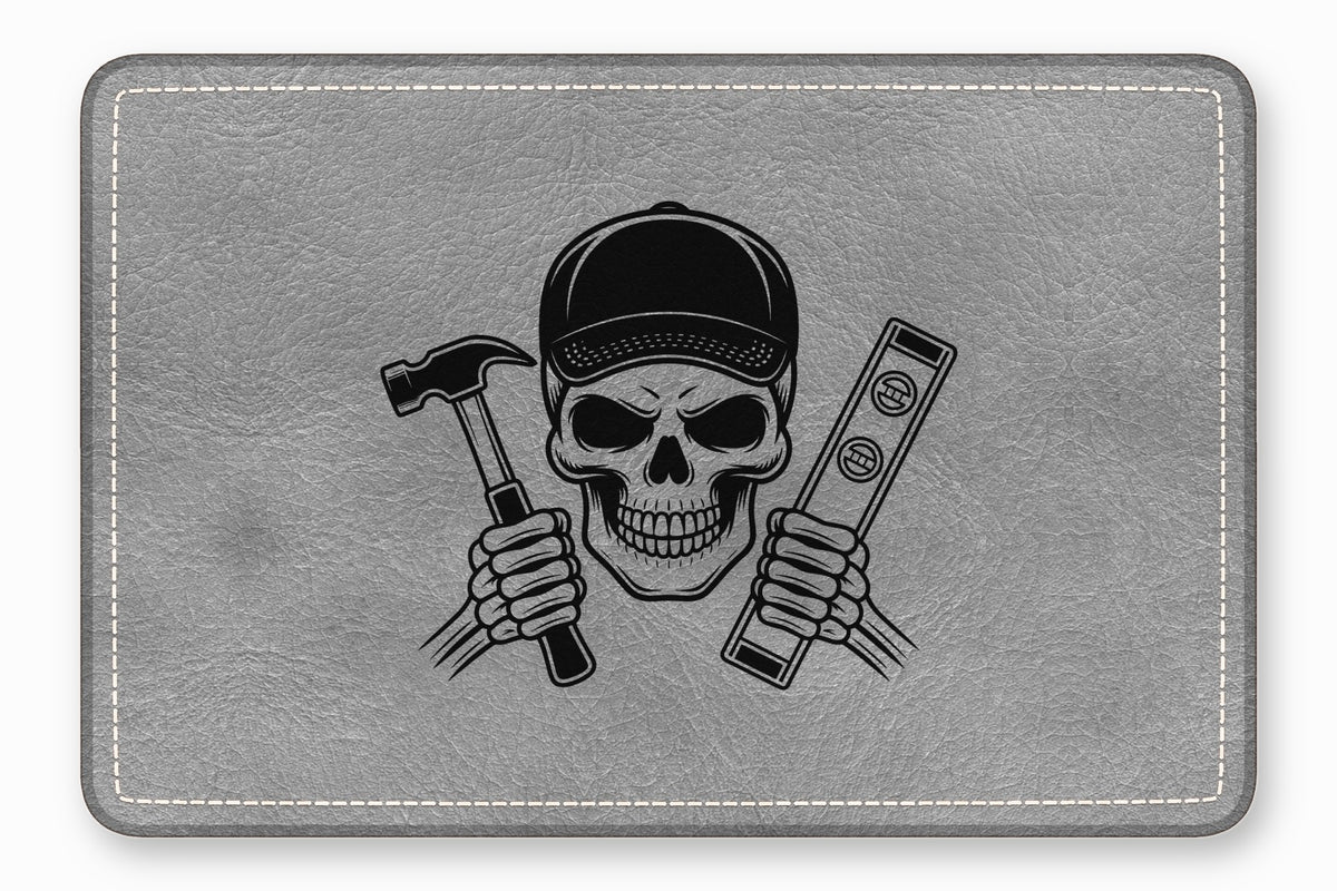 A rectangular grey leather patch featuring a skull wearing a cap and holding a hammer and level.