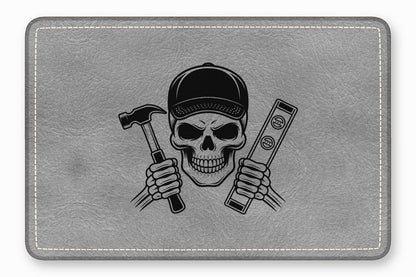 A rectangular grey leather patch featuring a skull wearing a cap and holding a hammer and level.