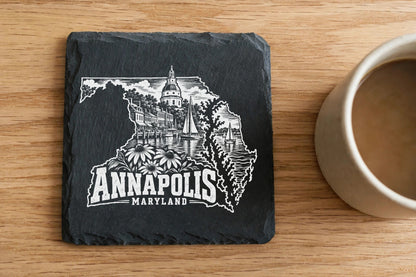 Square black slate coaster engraved with a white Annapolis, Maryland skyline design next to a coffee mug on wood.