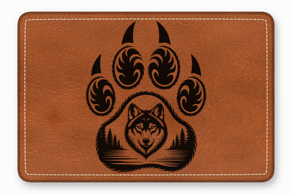 A rectangular brown leather patch features a laser-engraved design of a bear paw containing a wolf and forest.