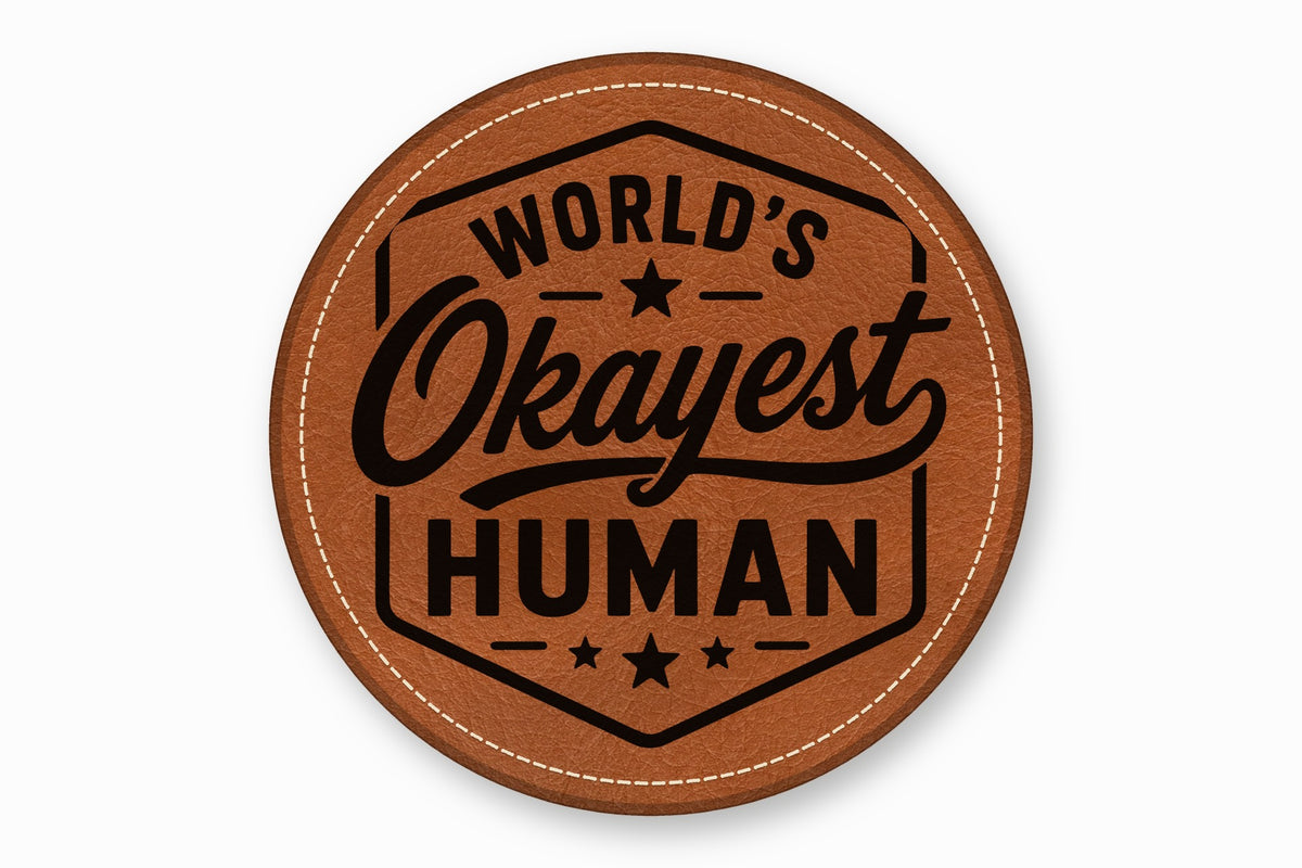 A round brown leatherette patch with white stitching and the engraved text World's Okayest Human.