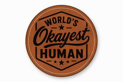 A round brown leatherette patch with white stitching and the engraved text World's Okayest Human.