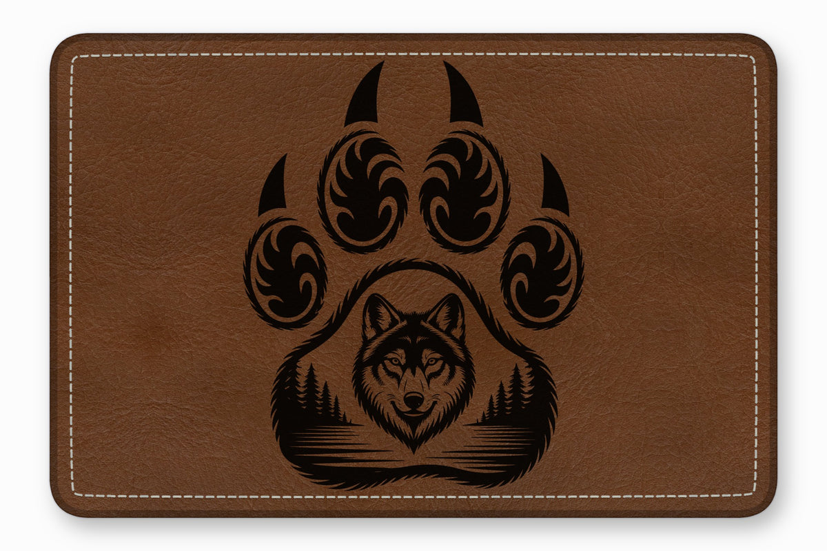 Brown leather patch with rounded corners featuring a black engraved bear paw enclosing a wolf head and forest landscape.