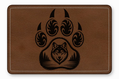 Brown leather patch with rounded corners featuring a black engraved bear paw enclosing a wolf head and forest landscape.