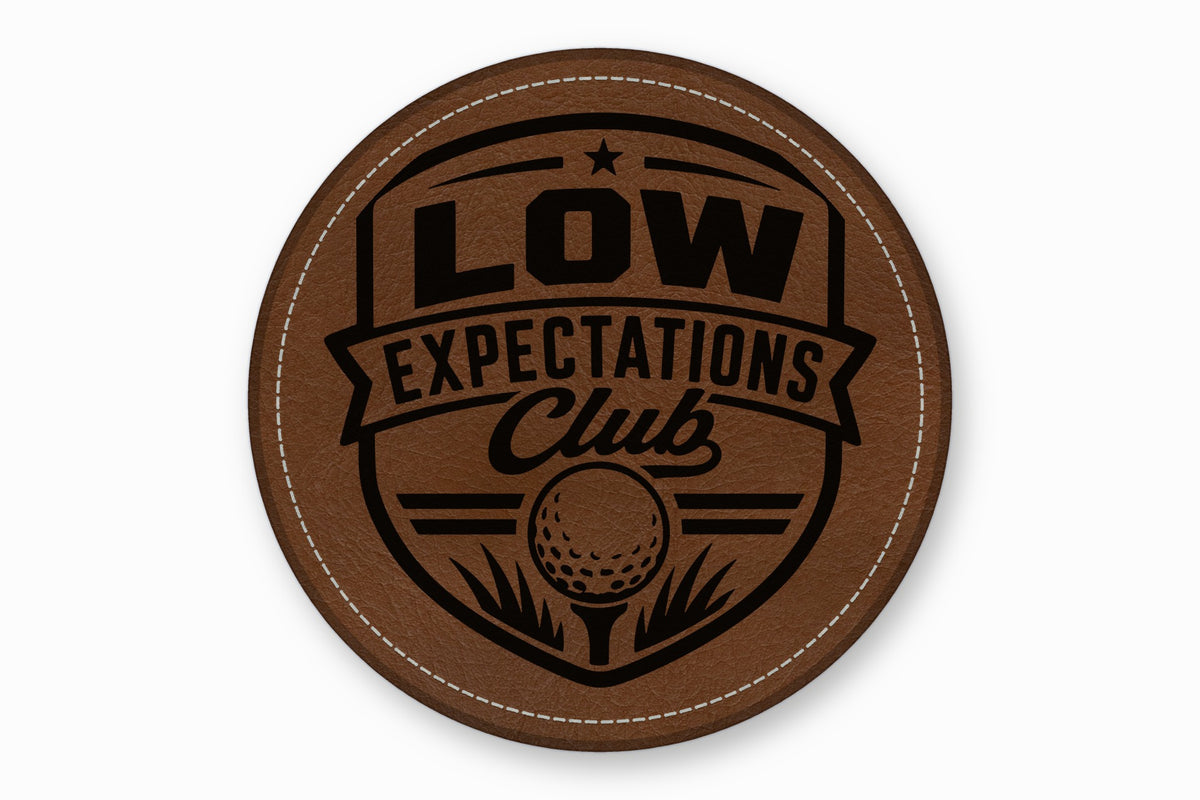 A round brown leatherette golf patch featuring a black engraved emblem and text reading "Low Expectations Club".