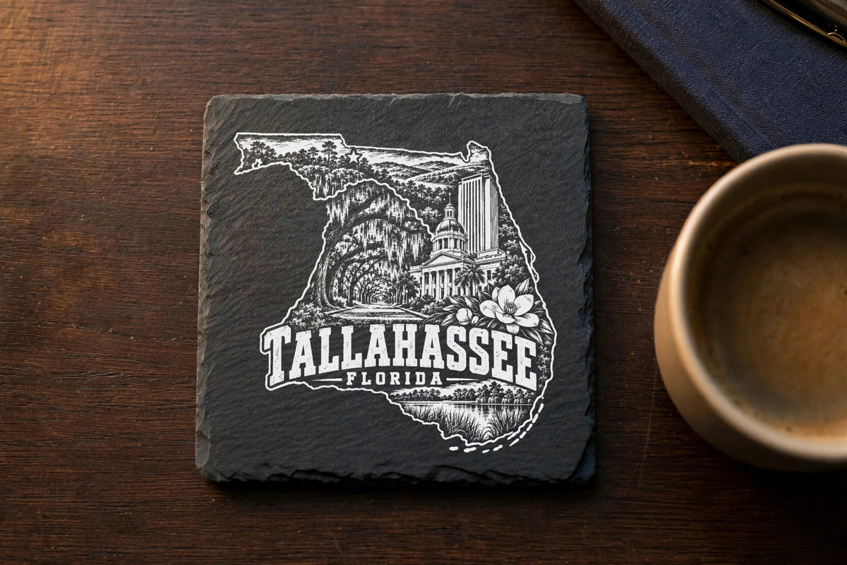 A square black slate coaster engraved with a Tallahassee, Florida map design sits on a wooden table.